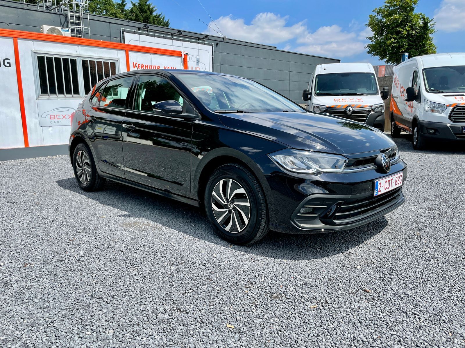 De zwarte Polo op de kiezel bij de loods, met de bestelwagens erachter